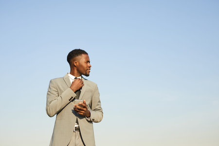 Content confident stylish young African-American businessman in beige suit standing against clear sky and adjusting necktie while using smartphoneの写真素材