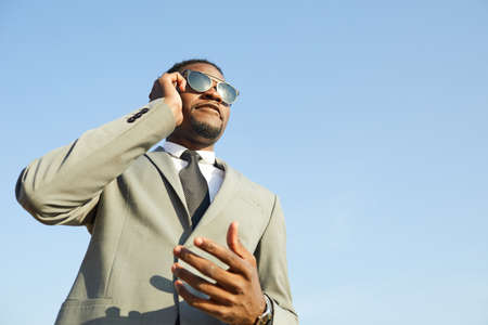 Below view of confident young black businessman with beard wearing stylish suit and sunglasses standing against blue sky and communicating on cellphoneの写真素材