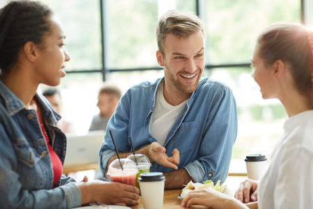 Group of jolly young multi-ethnic students in casual outfits leaning on table and eating fast food...