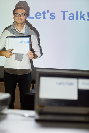 Serious young sales manager in eyeglasses standing in front of projection screen with text and...