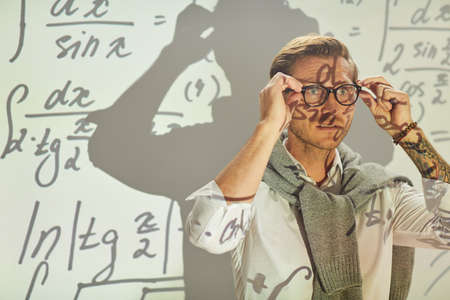 Serious pensive young math analyst with sweater around neck adjusting eyeglasses while preparing for solving equationsの写真素材