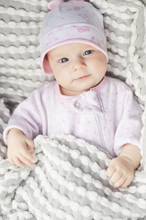 Sweet small baby with big blue eyes in white and pink costume and hat lying under comfy grey blanket