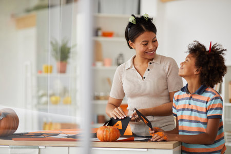 Cheerful young black mother and cute son with Afro hairstyle standing at counter and preparing paper decorations for Halloween with pleasureの写真素材