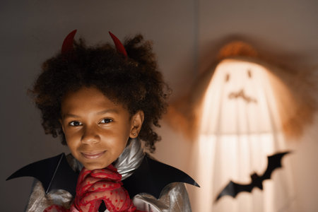 Portrait of dreamy cute Afro-American boy in Halloween dress leaning head on hands in red gloves in...