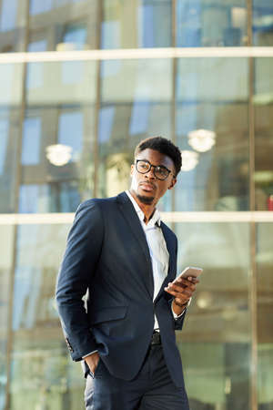 Thoughtful young African American businessman in suit and glasses standing against contemporary building and holding smart phoneの写真素材