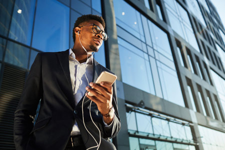 Below view of thoughtful young black businessman in earphone listening to voice message via phone app outdoorsの写真素材