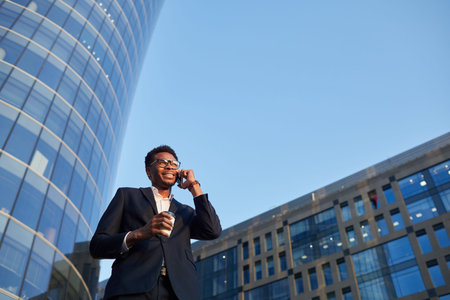 Content handsome young Afro-American manager in glasses walking over financial district and drinking...
