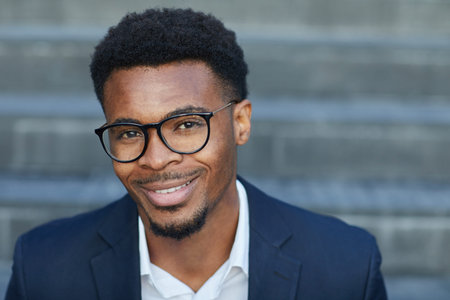 Headshot of positive handsome young black male manager with beard smiling at camera outdoors