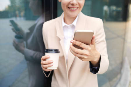 Close-up of positive lady in jacket standing by glassy building outdoors and texting via messenger on smartphoneの写真素材
