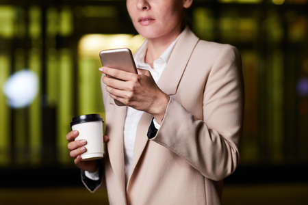 Close-up of thoughtful businesswoman in stylish jacket standing in dark office and drinking coffee while checking planner app on smartphoneの写真素材