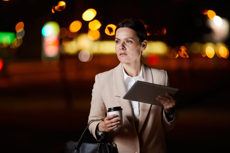 Serious introspective middle-aged businesswoman in jacket drinking coffee and using tablet while...