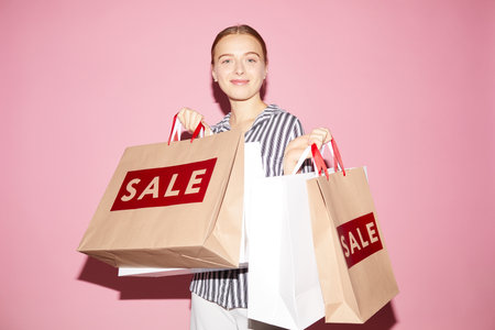 Portrait of smiling attractive young woman standing against pink wall and holding paper bags while enjoying big saleの写真素材