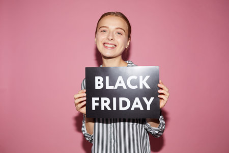 Cheerful beautiful young woman in stripped shirt standing against pink background and showing banner...