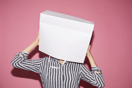 Anonymous woman in black and white shirt standing against pink wall and holding white paper bag over...