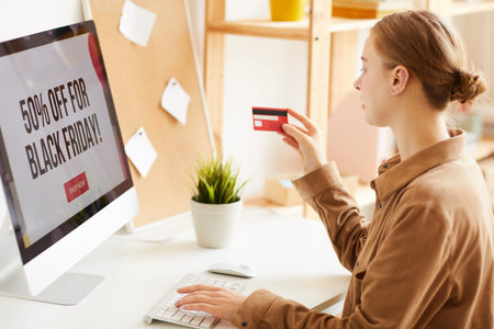 Young woman with hair bun sitting at desk and using credit card while paying online via website