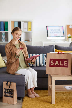 Smiling young woman in stylish outfit sitting on sofa at home and using tablet and credit card for online payment, paper bags with Sale and Black Friday inscriptionsの写真素材