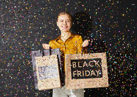 Jolly excited young woman in blouse standing under glittering confetti against black wall at Black...