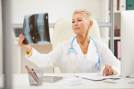 Pensive attractive mature radiologist with stethoscope on neck sitting at table and analyzing x-ray...