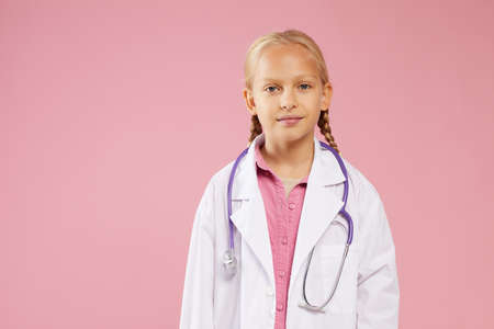 Content confident girl in lab coat wearing purple stethoscope around neck standing against pink background and looking at cameraの写真素材