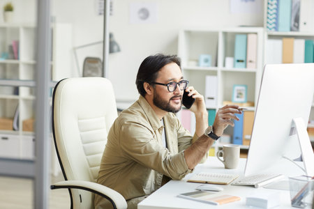 Smart young Asian programmer in casual shirt sitting at table with notepads and pointing at computer monitor while talking to customer by phoneの写真素材