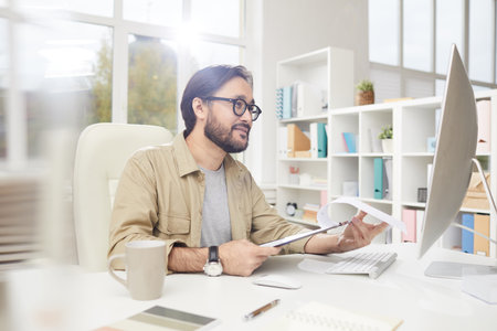 Content handsome hipster young Asian man with beard sitting at table and checking papers while talking to colleagues via video conferencing appの写真素材