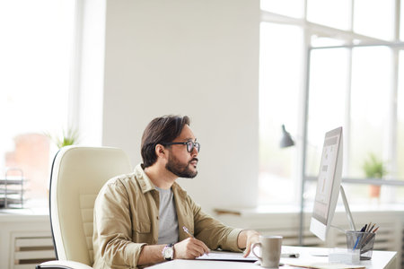 Serious hipster young Asian designer in glasses sitting at desk and looking at computer monitor...