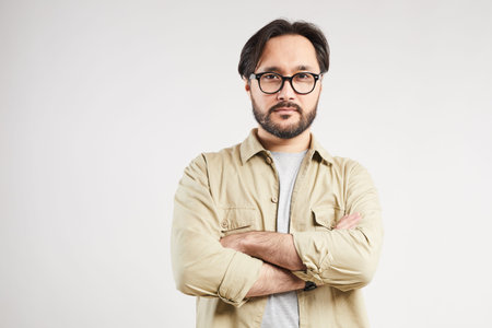 Portrait of serious handsome young Asian man in khaki shirt wearing stylish eyeglasses crossing arms...