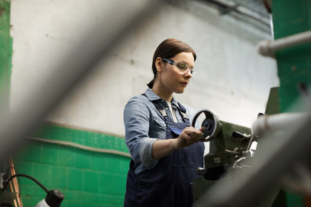 Serious middle-aged female factory worker in protective goggles using tailstock while processing...