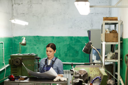 Thoughtful middle-aged female engineer in workwear standing at metal counter and reading technical...