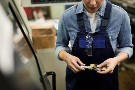 Close-up of serious female factory worker with safety goggles hanging on overall standing by...