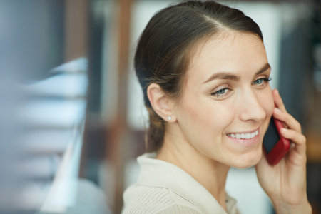 Smiling friendly young secretary with brown hair talking to customer by mobile phone, reflection...