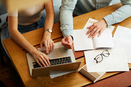 High angle view of unrecognizable colleagues sitting at wooden table in office and using laptop and papers while comparing data togetherの写真素材