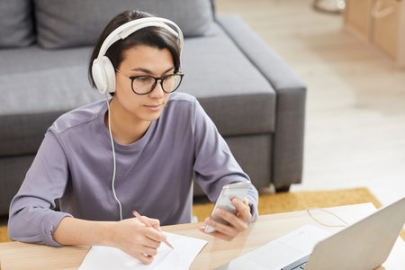 Pensive young freelance marketer in wired headphones and glasses sitting at coffee table and making...