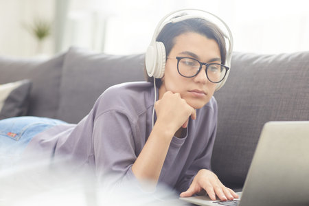 Serious pensive attractive girl in wired headphones lying on sofa and using laptop while viewing...
