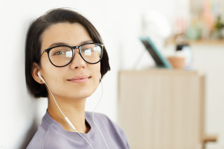 Portrait of smiling hipster girl in earphones and spectacles sitting by wall at home and listening to musicの写真素材
