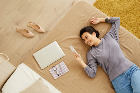 Directly above view of happy young woman with short hair lying on bed and enjoying online...
