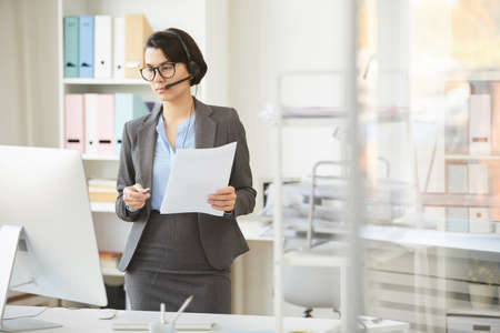 Serious young business lady in hands-free device standing at desk with computer and discussing project plan with team via video chatの写真素材