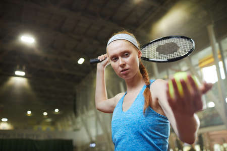 Serious focused female professional tennis player in blue shirt and headband standing on indoor court and serving ball while starting matchの写真素材