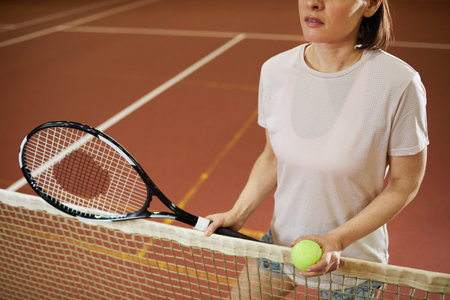 Close-up of content woman in comfortable tshirt standing by net and holding ball and racket while...