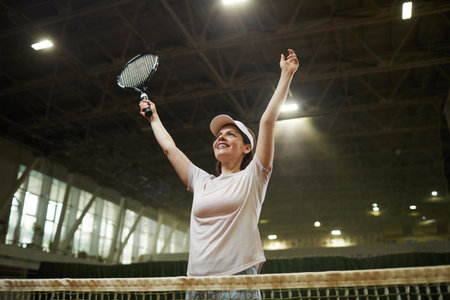 Cheerful excited young woman in cap standing by net and raising arms up while celebrating winning in tennis matchの写真素材
