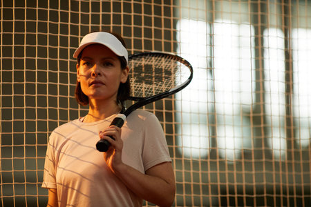 Portrait of serious confident female tennis player in cap standing against net on court and keeping...