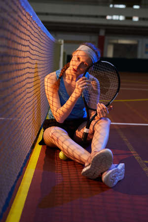 Portrait of serious pretty female tennis player in sports headband sitting on dark court floor and embracing racquetの写真素材