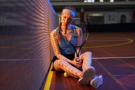 Portrait of serious beautiful young female tennis player with red braid sitting on floor at dark indoor court and holding racketの写真素材