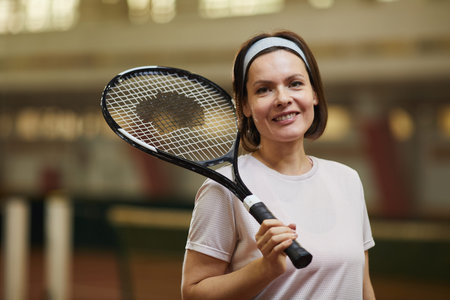 Portrait of happy energetic middle-aged woman in sports headband standing on court and holding...