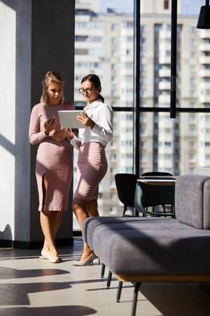 Serious thoughtful young businesswomen in pink clothes standing in modern hall and using tablet...