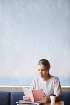 Serious attractive blond woman in sweater sitting at table with laptop and coffee cup and reading...