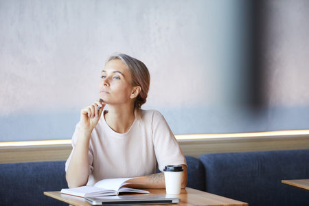 Serious young businesswoman with tattoo on arm sitting at table in coffee shop and thinking of new...