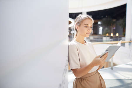 Serious pensive young woman with ponytail leaning on wall in lobby and reading interesting bookの写真素材