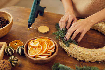 Close-up of unrecognizable woman with manicure standing at wooden table in workshop and using hot glue while attaching fir tree twigs to wreath baseの写真素材