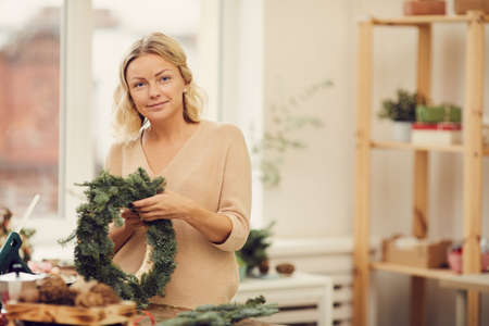 Portrait of smiling attractive young blonde woman in sweater standing at table in workshop and adding tree twigs to wreathの写真素材